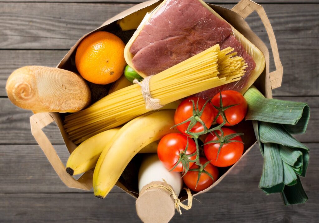 Full paper bag of various groceries on wooden background, top view