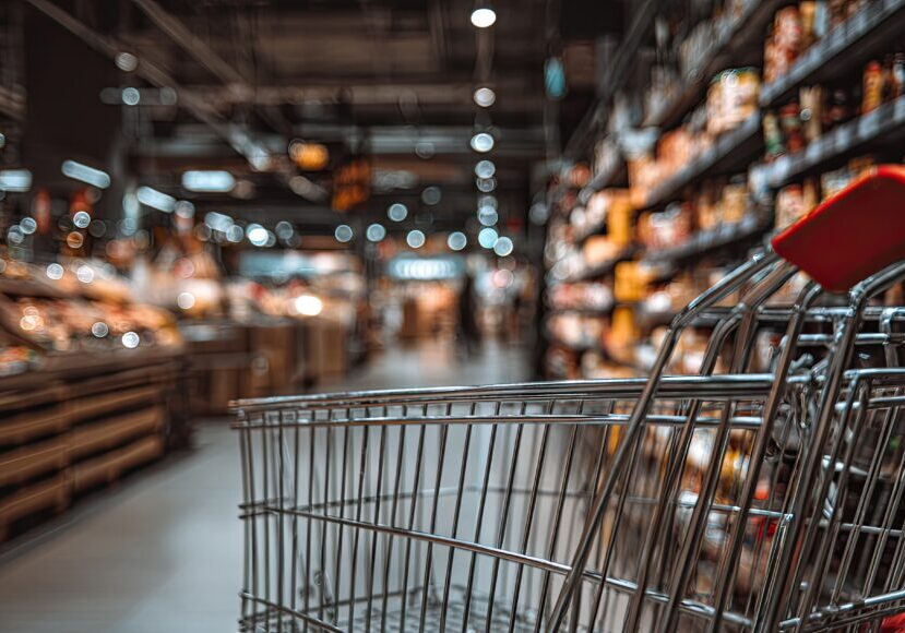 Shopping cart is centrally situated in a dimly lit grocery store aisle, with the background exhibiting a soft focus effect on various food items and shelves.  The cart's metallic frame and red handles are in sharp focus, while the shelves and products in the background appear out-of-focus, providing a sense of depth and space.