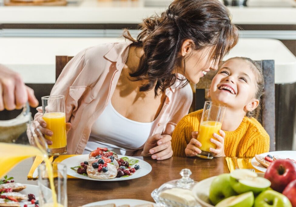 cheerful mother with daughter sitting at table with fresh juice and pancakes with berries at kitchen