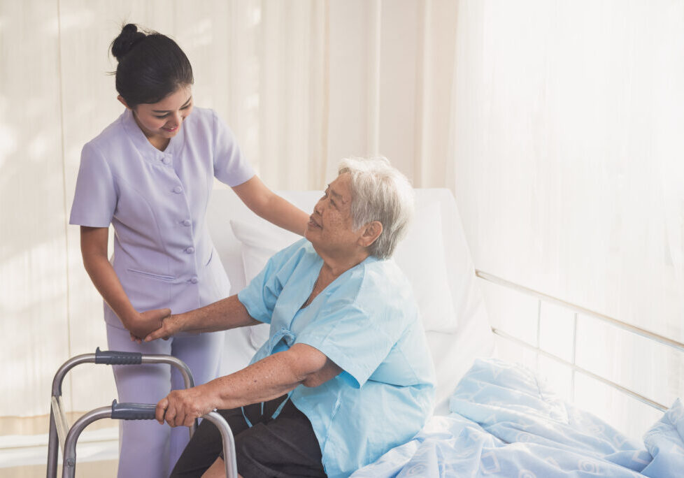 woman caregiver and elderly patient on examination couch. Happy nurse holding hand of senior to help senior patient. female nurse supporting senior disabled woman with walker at hospital. Health care and people concept