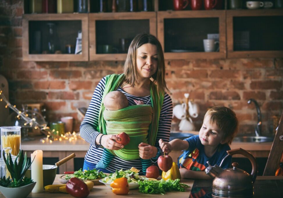 Happy young family, beautiful mother with two children, adorable preschool boy and baby in sling cooking together in a sunny kitchen. Vintage style.