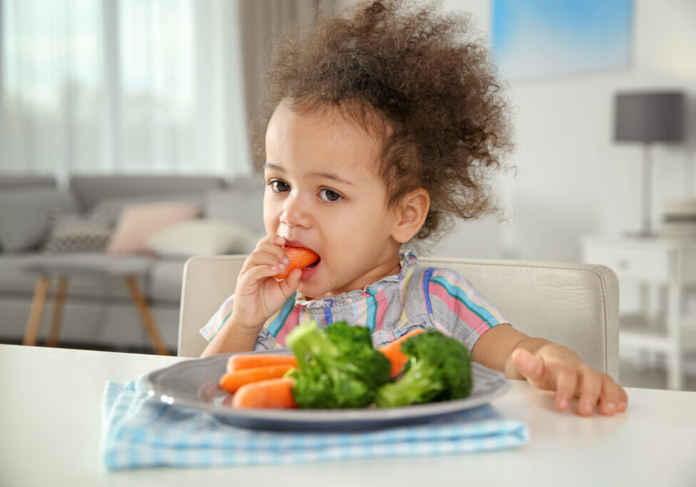 Cute African-American girl eating vegetables at table in living room