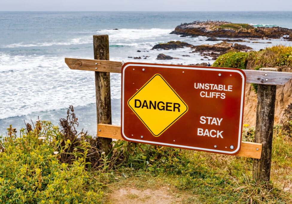 "Danger, unstable cliffs, stay back" sign posted on the Pacific Ocean coastline on an area with eroded cliffs and risk of landslide, California