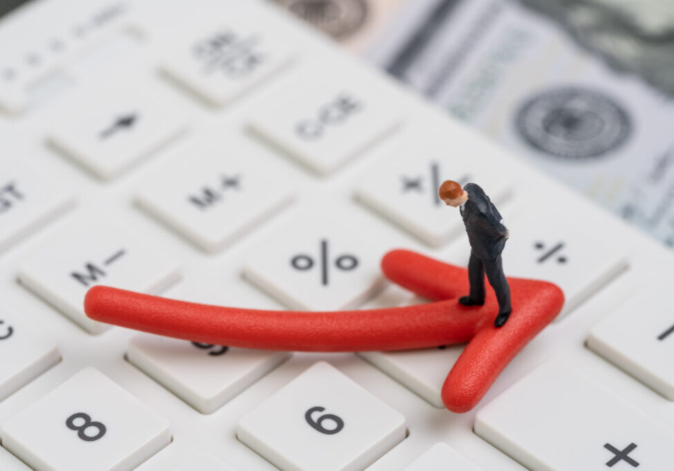 Economic recession, bear stock market or financial crisis concept, miniature businessman standing on red pointing down arrow on white calculator with background of US dollar banknote money.