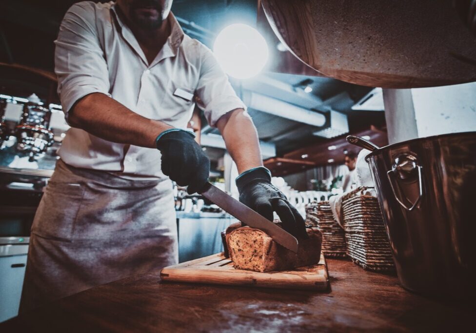 Expirienced baker in protective gloves is slicing bread for daily breakfast at restaurant.