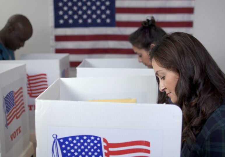 Side view MS of three people, two women and one man, Caucasian, Hispanic and African American, voting in booths at polling station. Large US flag on wall behind