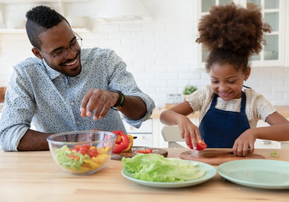 Smiling young african American father and small ethnic daughter chop vegetable cook healthy vegetarian salad together, happy biracial dad and little girl child have fun preparing food in home kitchen