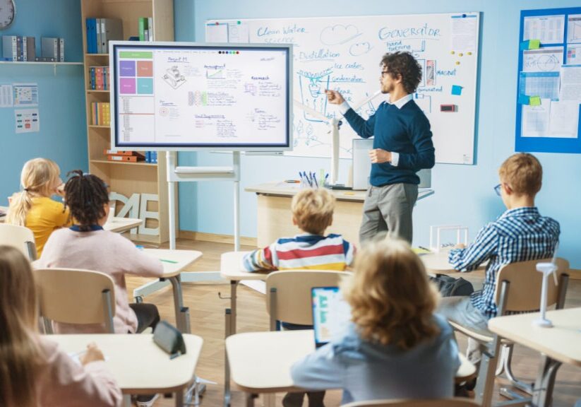 Elementary School Science Teacher Uses Interactive Digital Whiteboard to Show Classroom Full of Children how Software Programming works for Robotics. Science Class, Curious Kids Listening Attentively