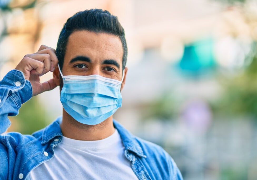 Young hispanic man putting on medical mask standing at the city.