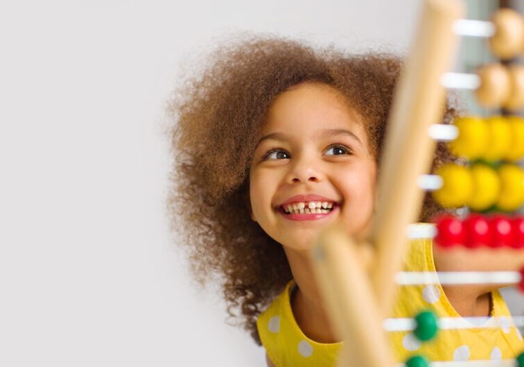 A Black student in a yellow dress laughs brightly behind a colored abacus in an elementary school classroom