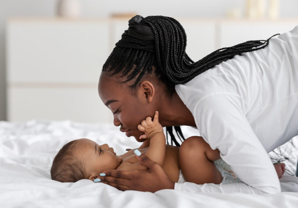 Happy Motherhood Concept. Closeup profile portrait of cheerful young black woman playing with her adorable small baby who lying in bed on white bedsheets, kissing sweet infant child
