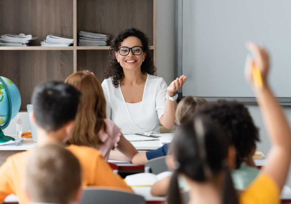 positive african american teacher pointing at blurred girl raising hand in classroom