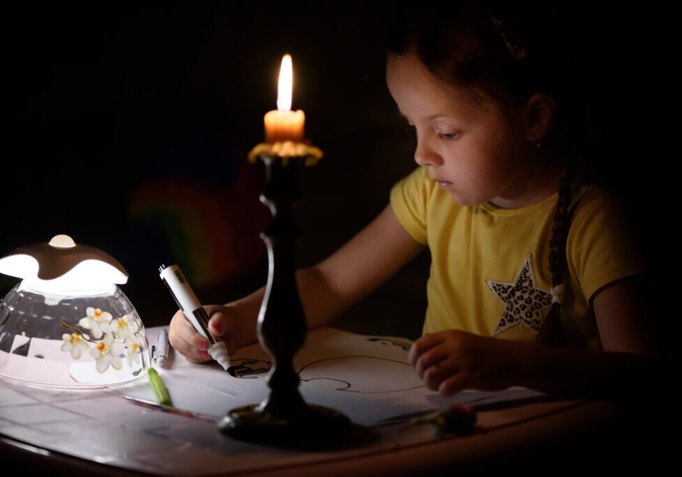 Little girl draws fantasy characters near candle in a dark room. Child doing favorite thing during a power outage.