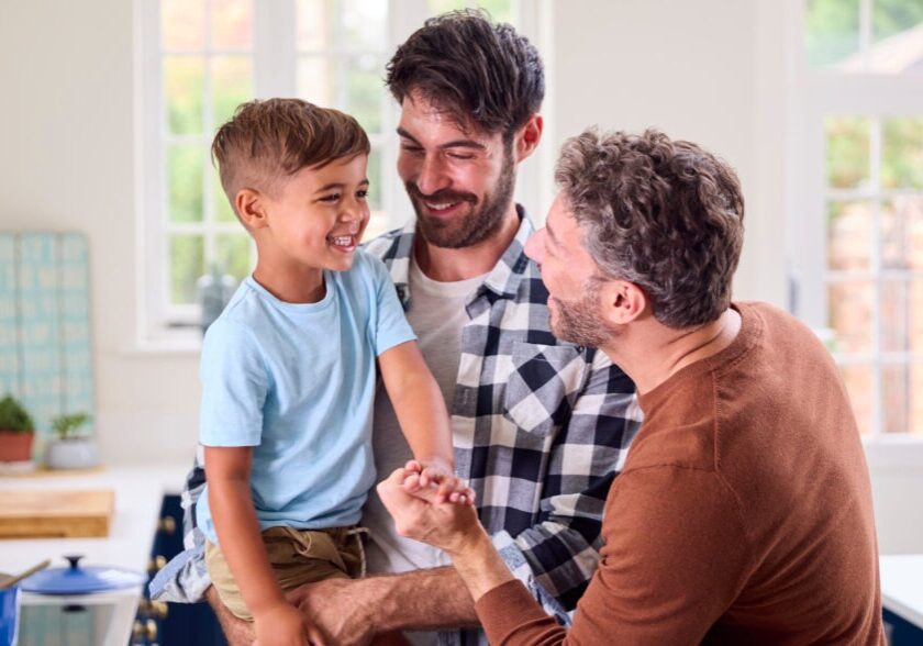 Same Sex Family With Two Dads Cooking In Kitchen With Son Sitting On Counter